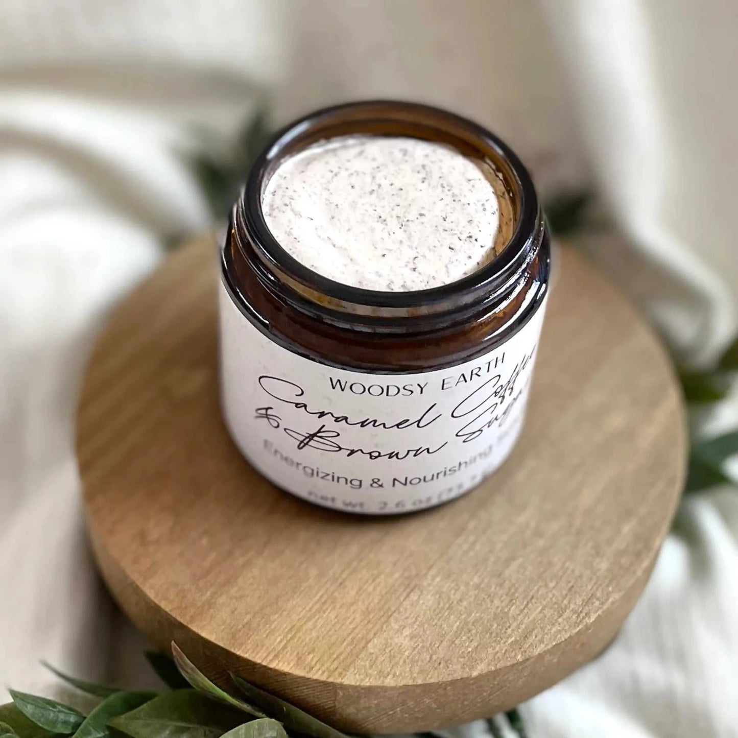 Open jar of Woodsy Earth Caramel Coffee Brown Sugar Scrub on wooden stand with greenery, natural exfoliating skincare.