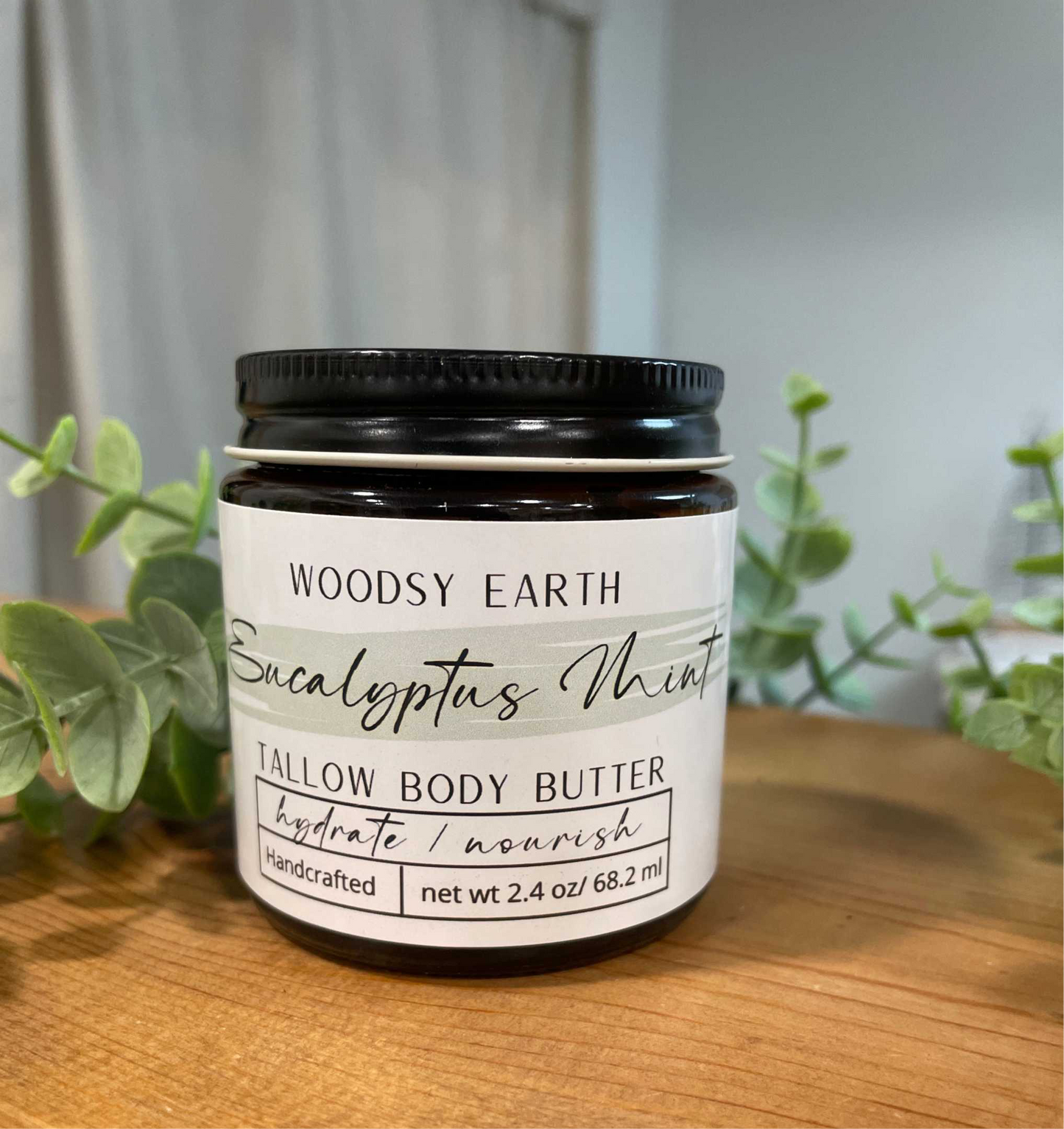 Woodsy Earth Eucalyptus Mint Tallow Body Butter in amber glass jar on wooden surface with eucalyptus leaves in the background