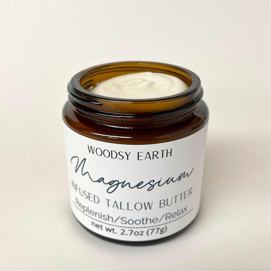An open amber glass jar of Woodsy Earth Magnesium Infused Tallow Butter, showing the white cream inside, set against a plain off-white background.