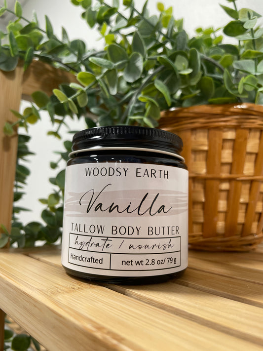 Vanilla Body Butter in a clear glass jar on wooden shelf with greenery in the background.