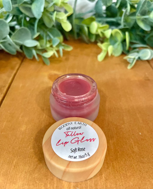 Tinted Tallow Lip Gloss in Soft Rose shade, open jar on wooden surface with greenery background.