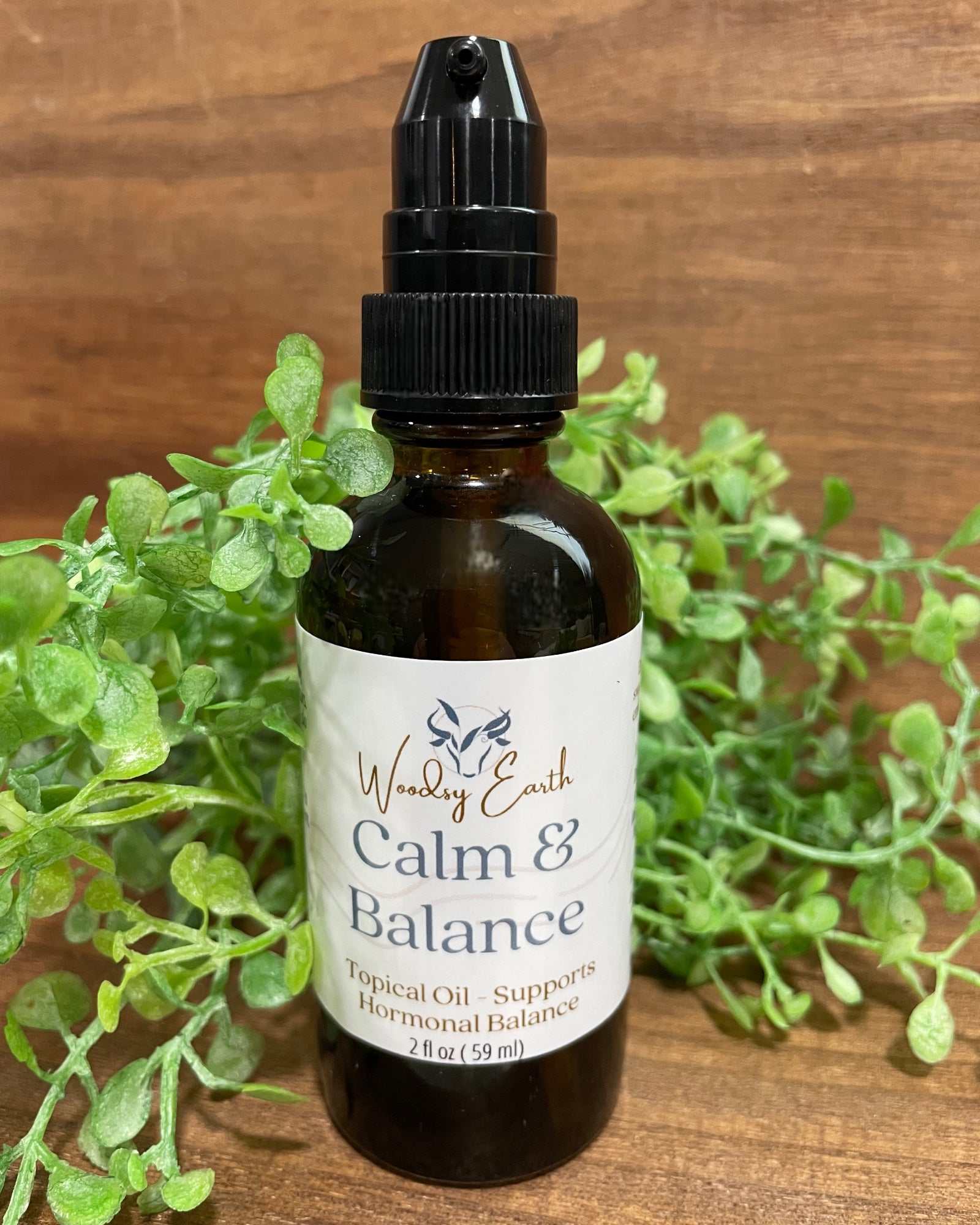 Woodsy Earth Calm & Balance topical oil in amber glass bottle with pump, labeled for hormonal support, displayed with greenery on wooden background.