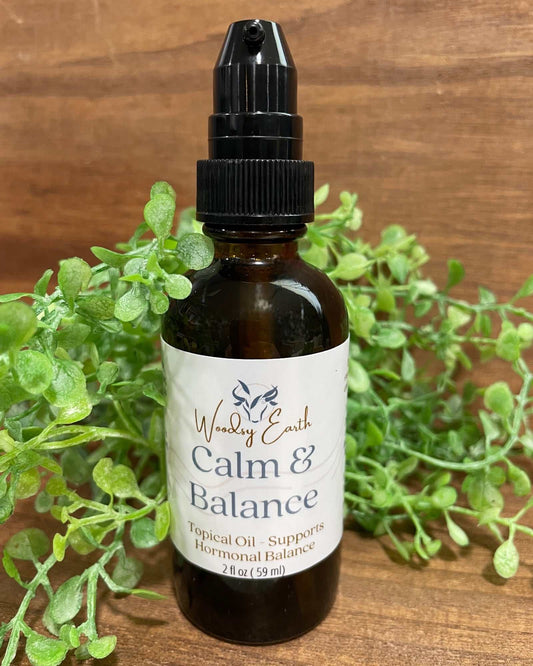 Woodsy Earth Calm & Balance topical oil in amber glass bottle with pump, labeled for hormonal support, displayed with greenery on wooden background.