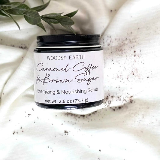 Woodsy Earth Caramel Coffee Brown Sugar Scrub jar on white cloth with greenery and coffee grounds, natural exfoliating skincare.