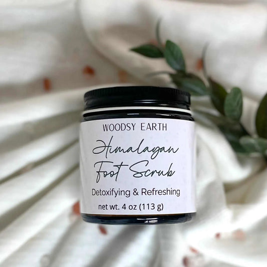 Woodsy Earth Himalayan Foot Scrub jar with greenery on white cloth, detoxifying and refreshing natural skincare.