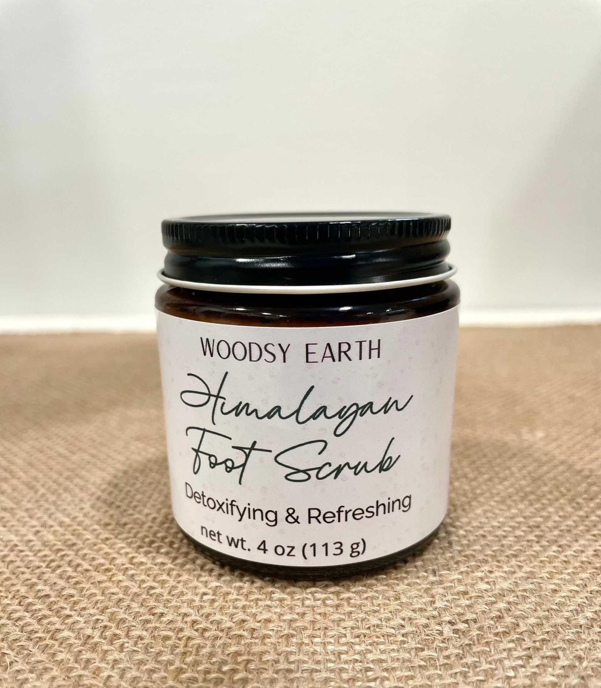 Woodsy Earth Himalayan Foot Scrub in amber glass jar on burlap surface, labeled detoxifying and refreshing