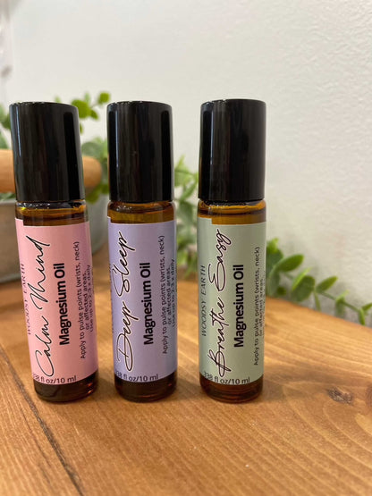 Woodsy Earth Everyday Balance collection of magnesium oil roll-ons in Calm Mind, Deep Sleep, and Breathe Easy blends, displayed in amber glass bottles on a wooden surface.