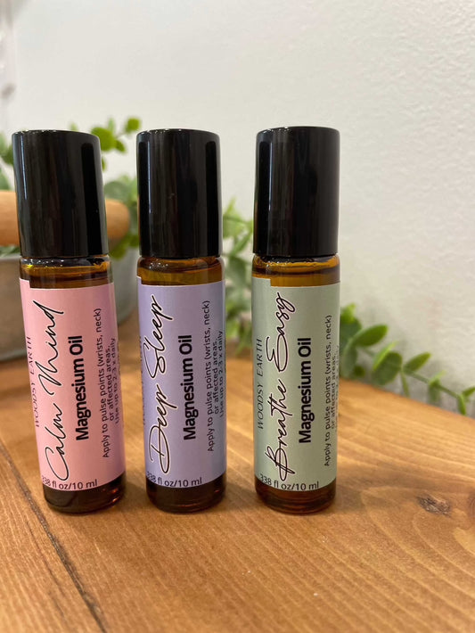 Woodsy Earth Everyday Balance collection of magnesium oil roll-ons in Calm Mind, Deep Sleep, and Breathe Easy blends, displayed in amber glass bottles on a wooden surface.