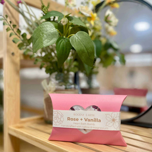 Pink bath bomb packaging with 'Rose + Vanilla' label on a wooden surface, blurred floral background