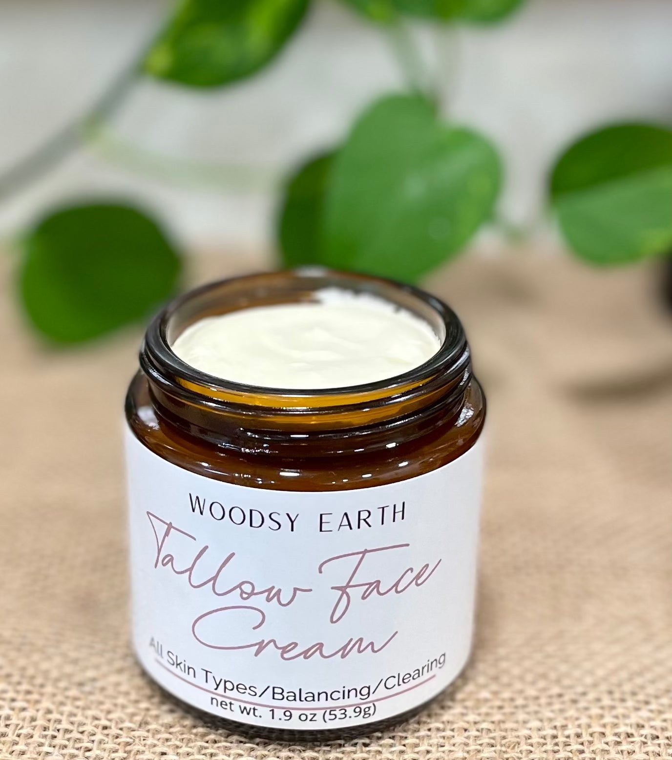 Open jar of Woodsy Earth Tallow Face Cream, 1.9 oz, on burlap with greenery in background