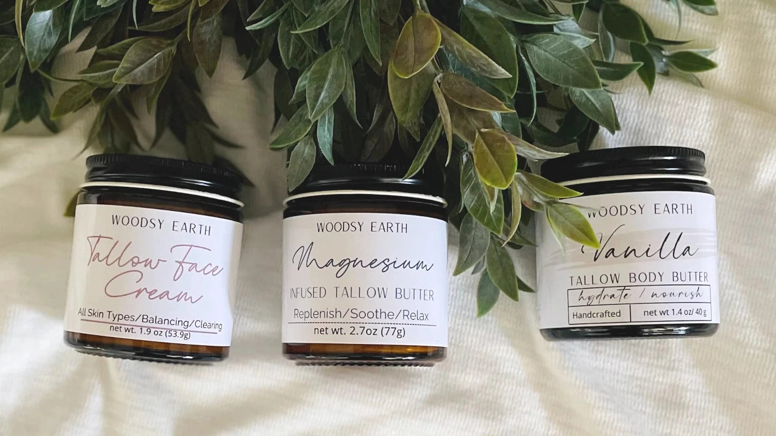Woodsy Earth natural skincare products – handcrafted tallow face cream, magnesium body butter, and vanilla tallow body butter displayed on fabric with greenery.