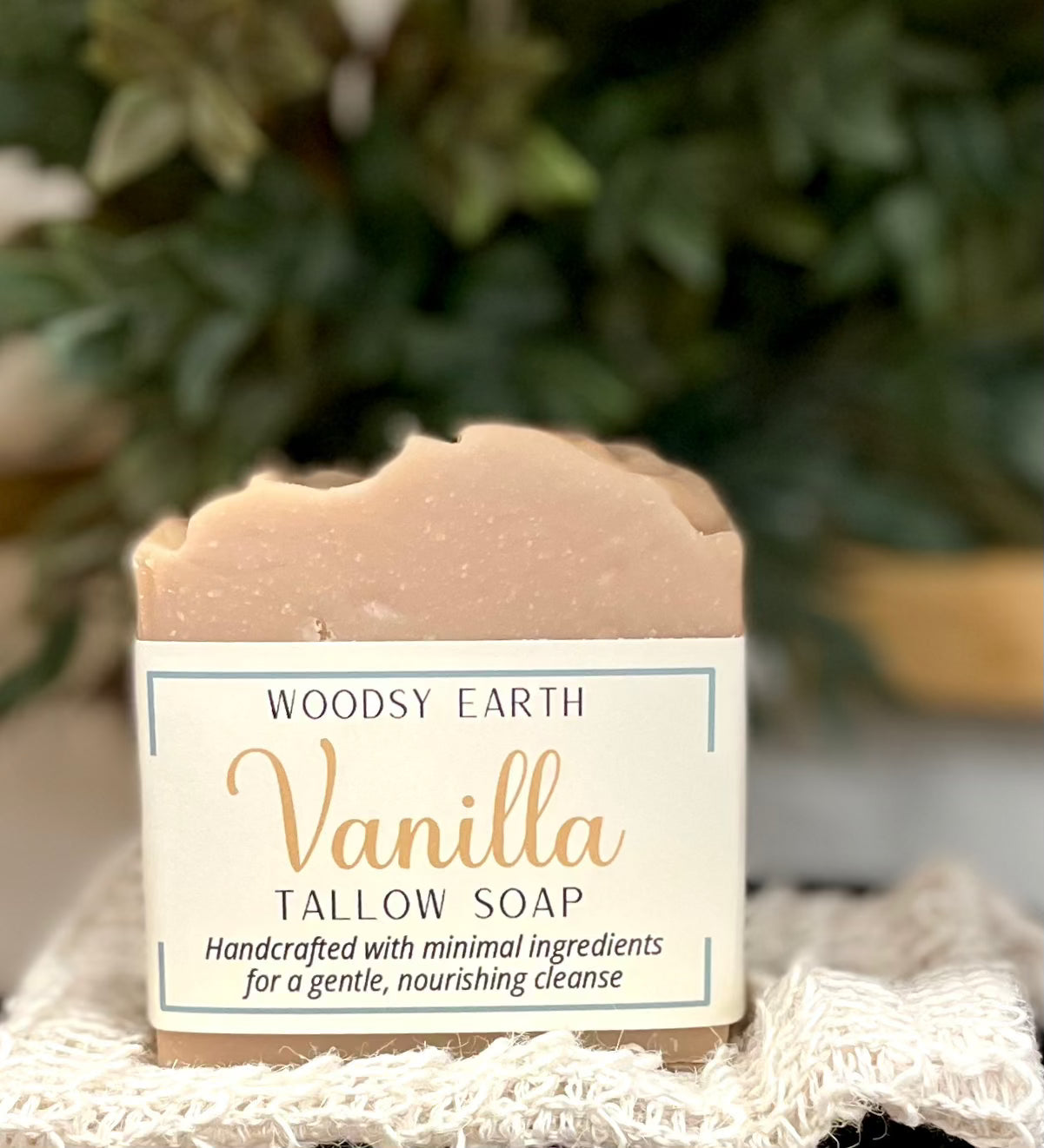 Woodsy Earth Vanilla Tallow Soap bar with creamy beige color, displayed on textured cloth with greenery background
